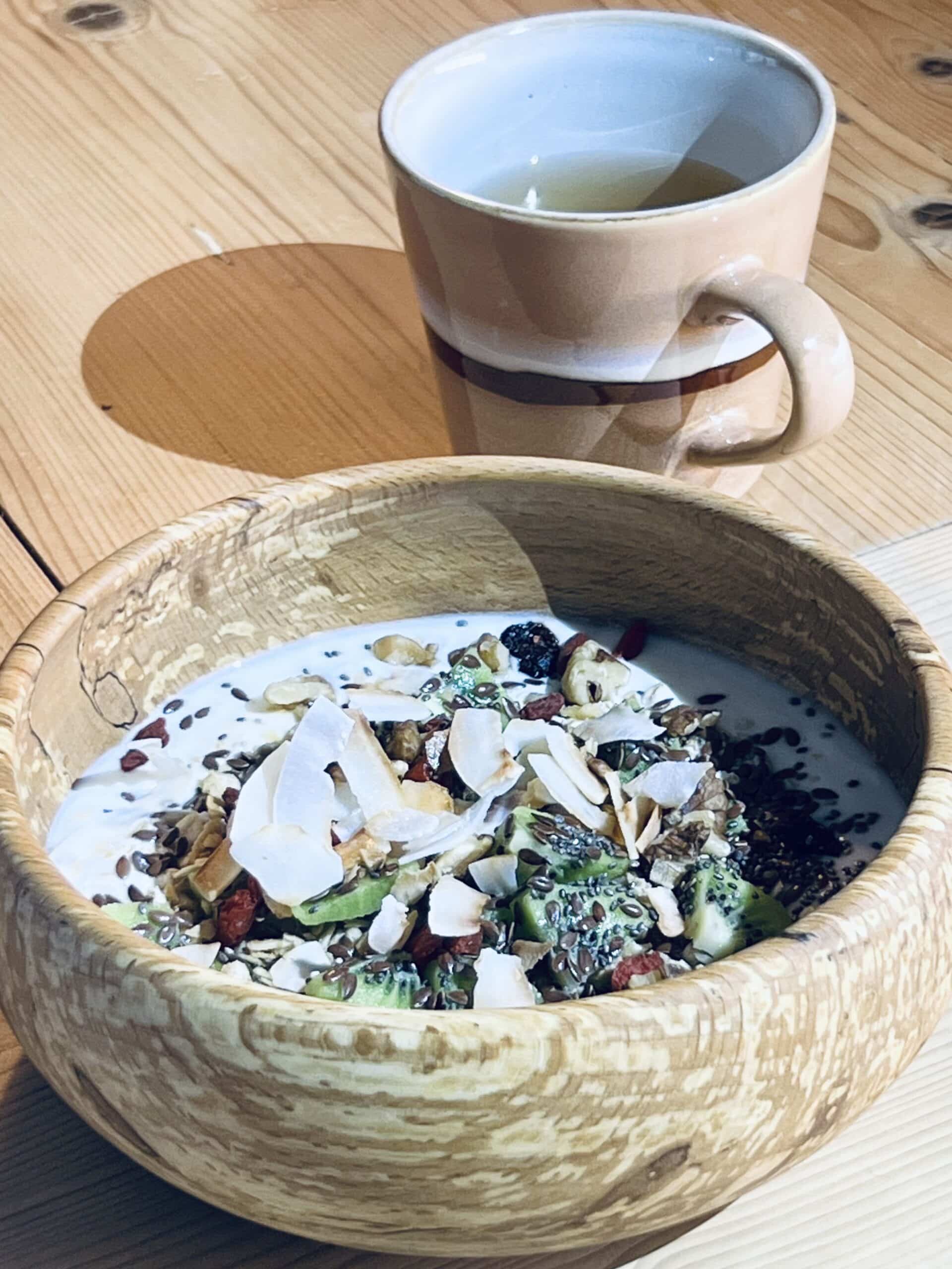 Een houten kom met yoghurt en ontbijtgranen, met een kop thee. Houten kom van het merk More than Wood. Salt + More Than Woods